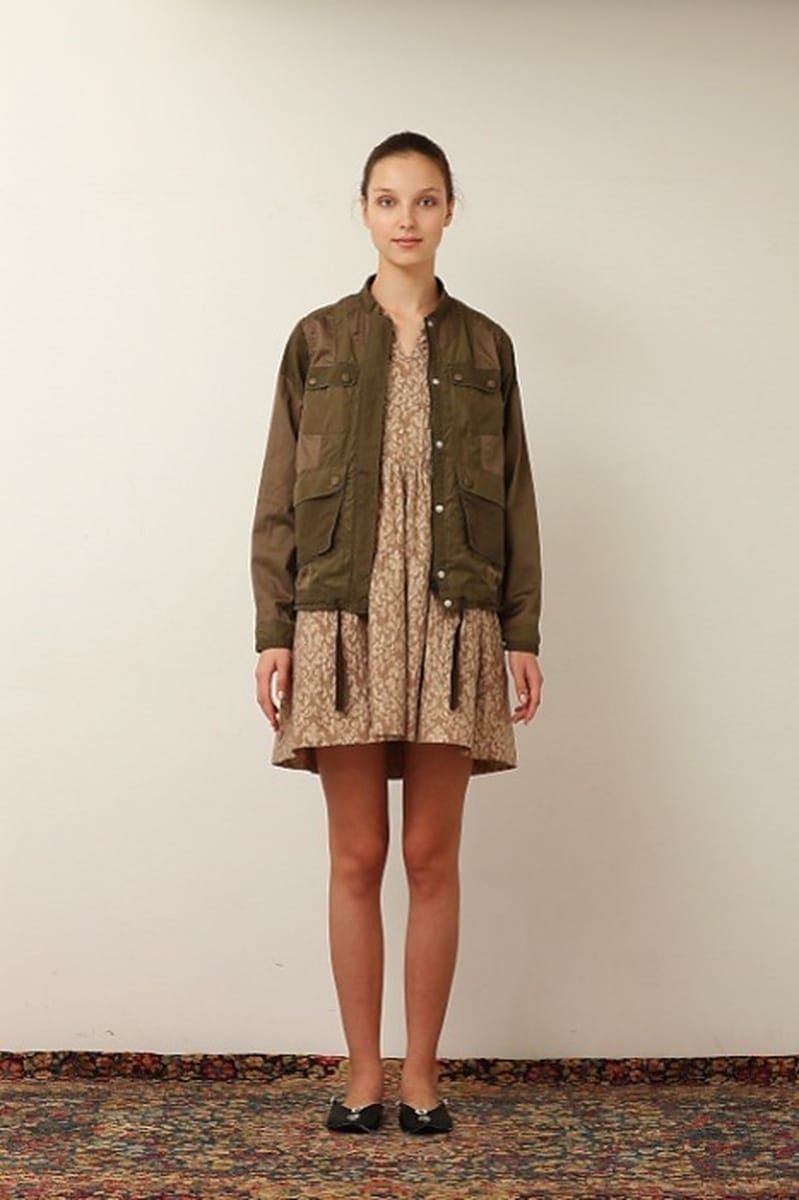 White Mountaineering women model in an olive jacket over a patterned dress, photographed against a plain studio backdrop.