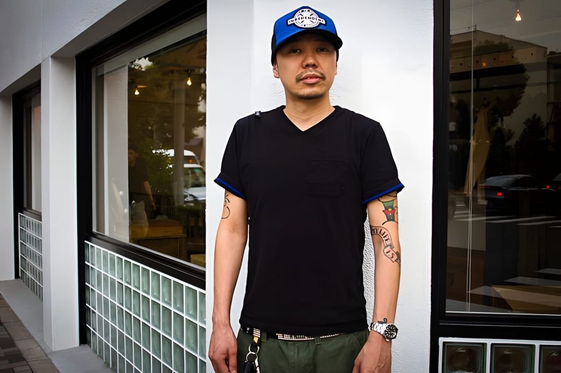 Tetsuya Nishiyama in blue Independent Trucks snapback, black v-neck tee, and olive trousers, standing outside a shop.