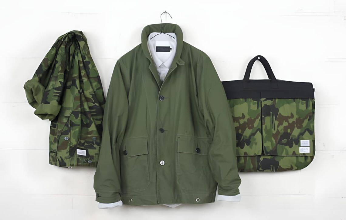 Maiden Noir olive field jacket on a hanger against a white wall, flanked by a camouflage jacket draped on the left and a camo