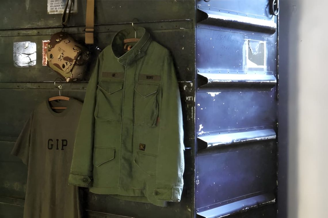 WTAPS GIP store interior: olive drab M-65 jacket and GIP logo tee on hangers, military helmet on distressed metal wall.