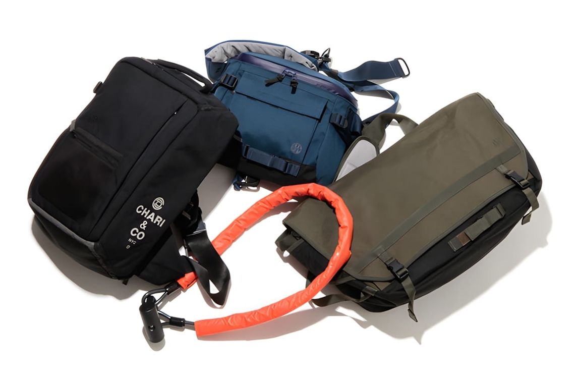 Three bags grouped together: black square Chari & Co NYC pack, blue sling, and olive messenger with orange padded strap in fo