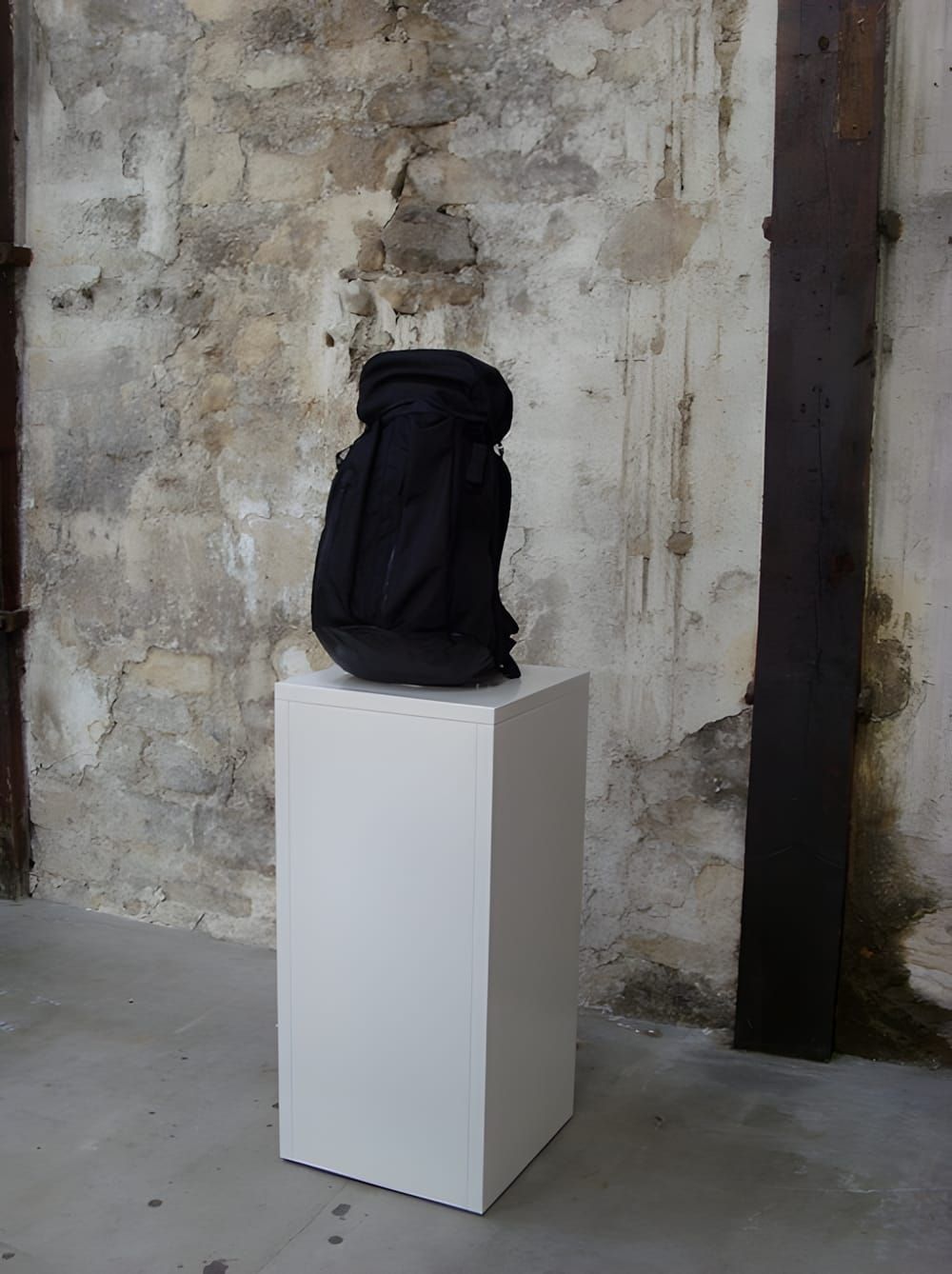 Black Visvim Ballistic 23L Cordura backpack displayed on white pedestal against raw plaster wall