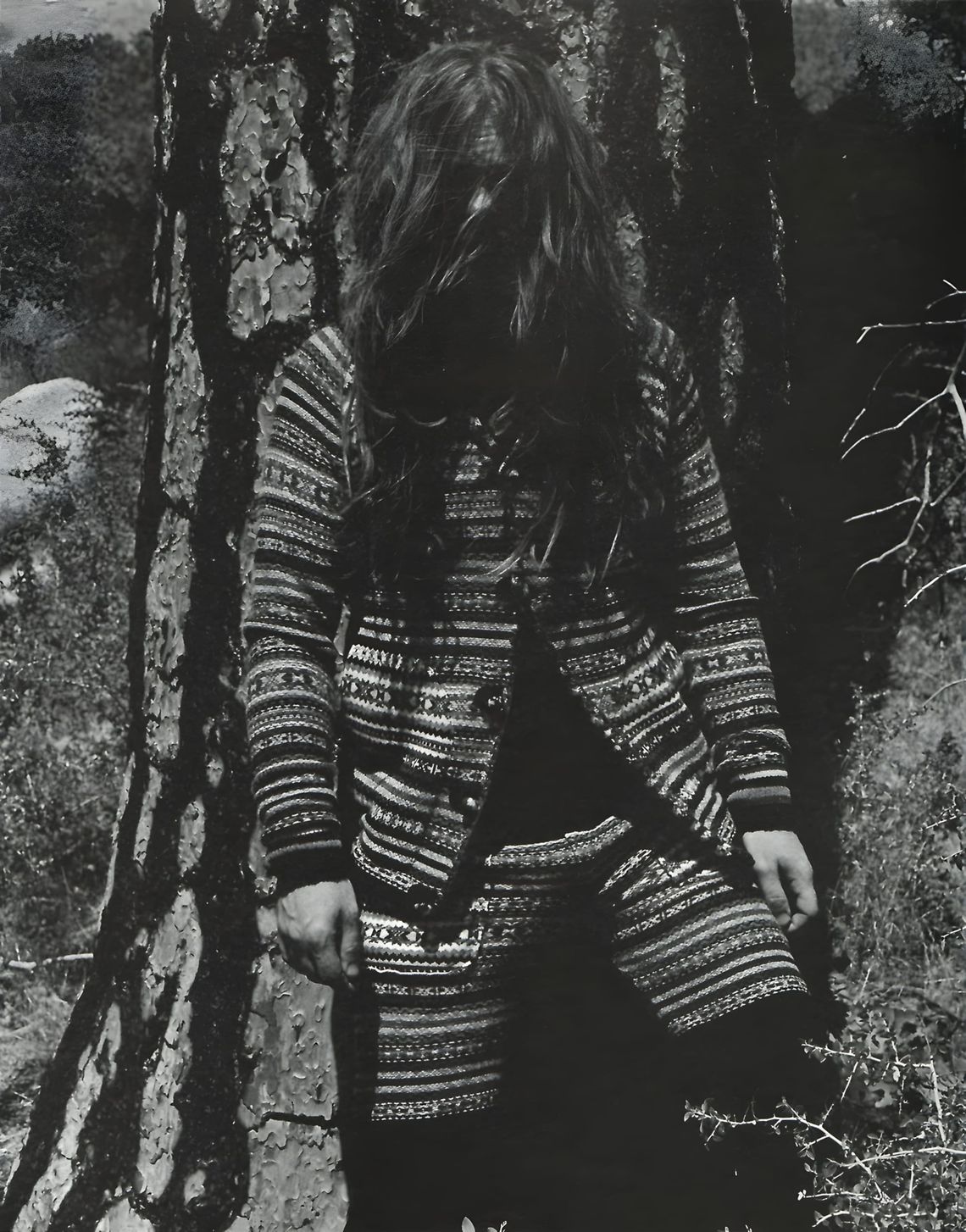 Figure in long patterned ethnic jacquard knit coat, face obscured by hair, standing by a tree — B&W editorial.