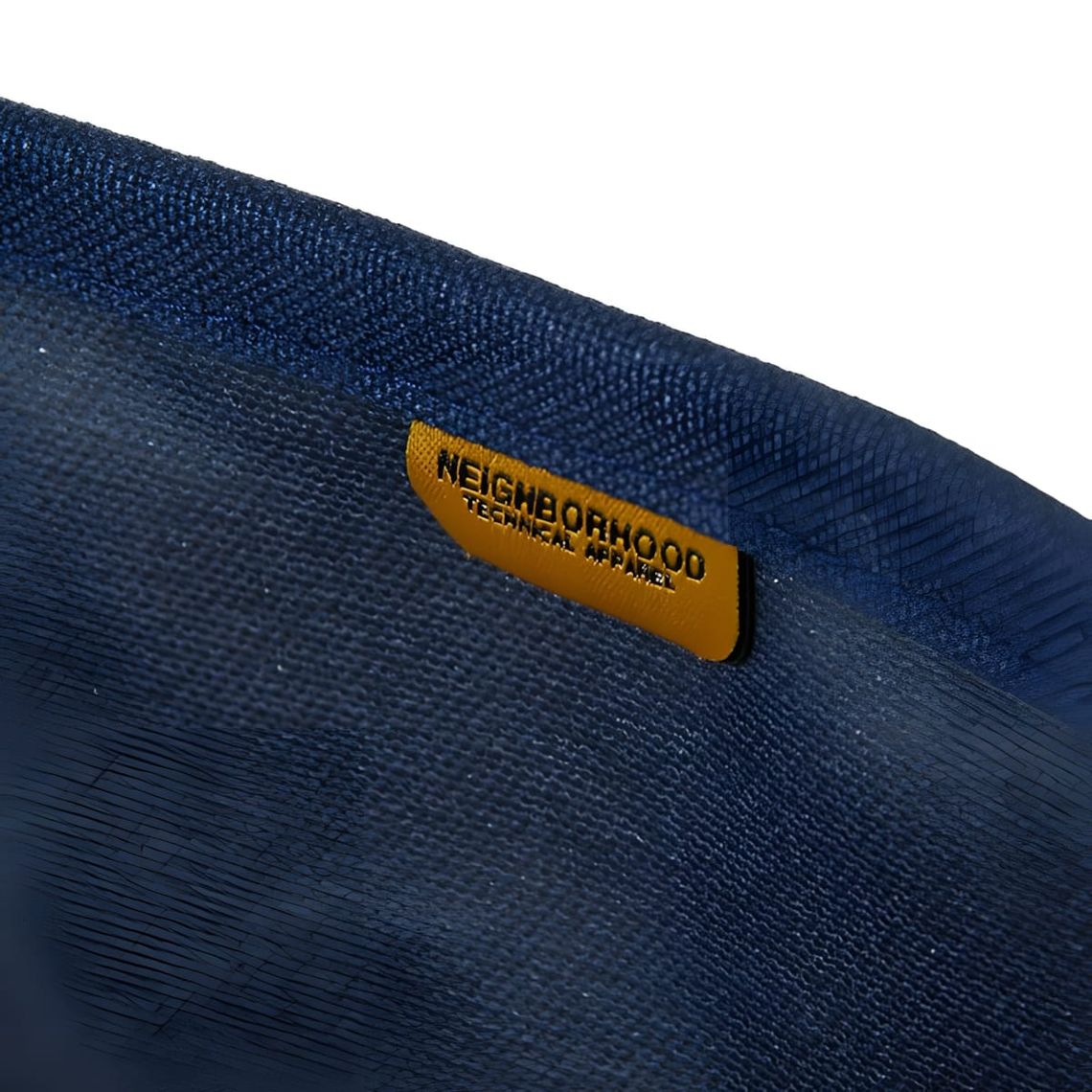 Close-up of an indigo selvedge denim pouch with a mustard amber leather tab reading 'Neighborhood Technical Apparel'