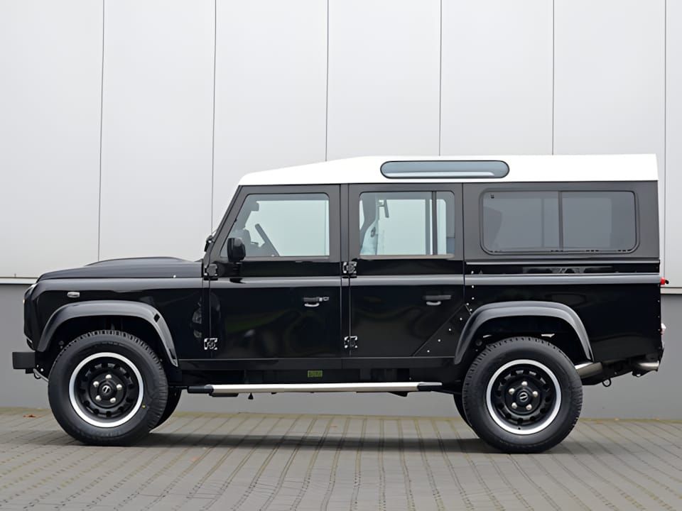 Land Rover Defender 110 Station Wagon in black with white roof, black steel wheels, profile view against grey warehouse wall.