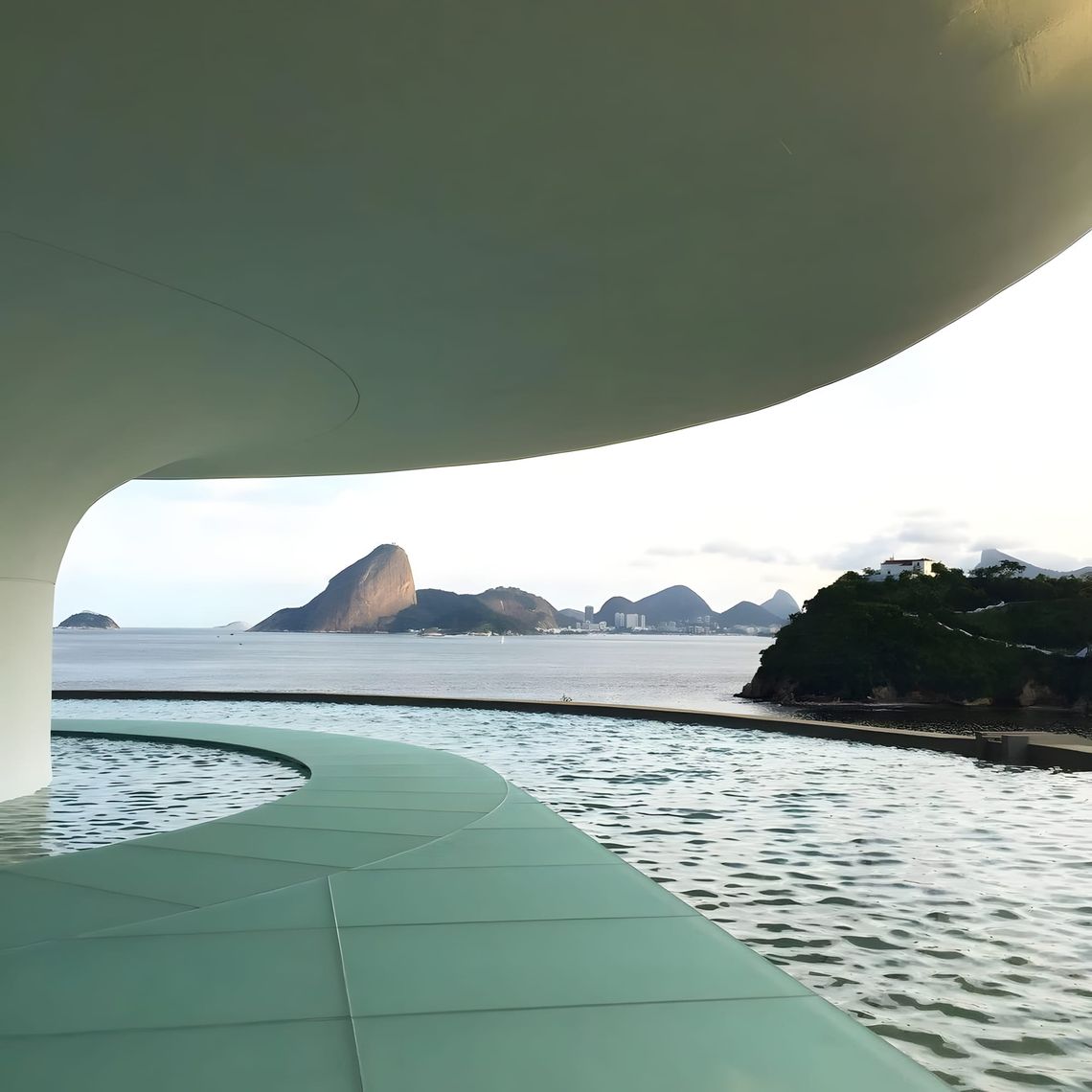 View from beneath the curved canopy of Oscar Niemeyer's MAC Niterói (1996): the mint-green ramp curves left into the reflecti