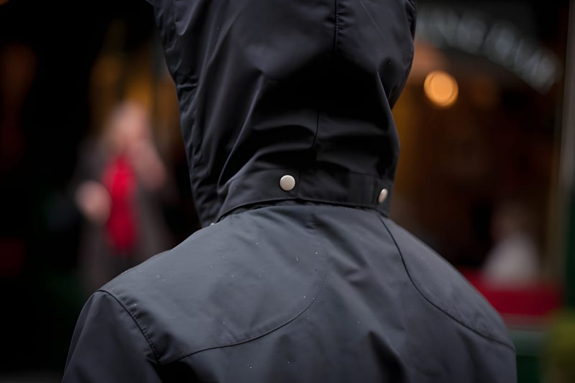 Back view of Folk SS2012 cotton rainmac in navy, collar buttoned up, silver snap hardware visible, urban background.