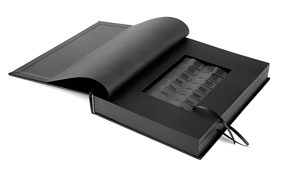 Matte black clamshell book box open to reveal a grid of cast bronze domino tiles in a cutout recess, with a black ribbon.