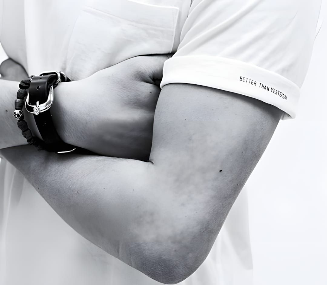 Crossed arms in WTAPS white tee with "Better Than Yesterday" sleeve-cuff print; beaded and buckle bracelets on wrist.