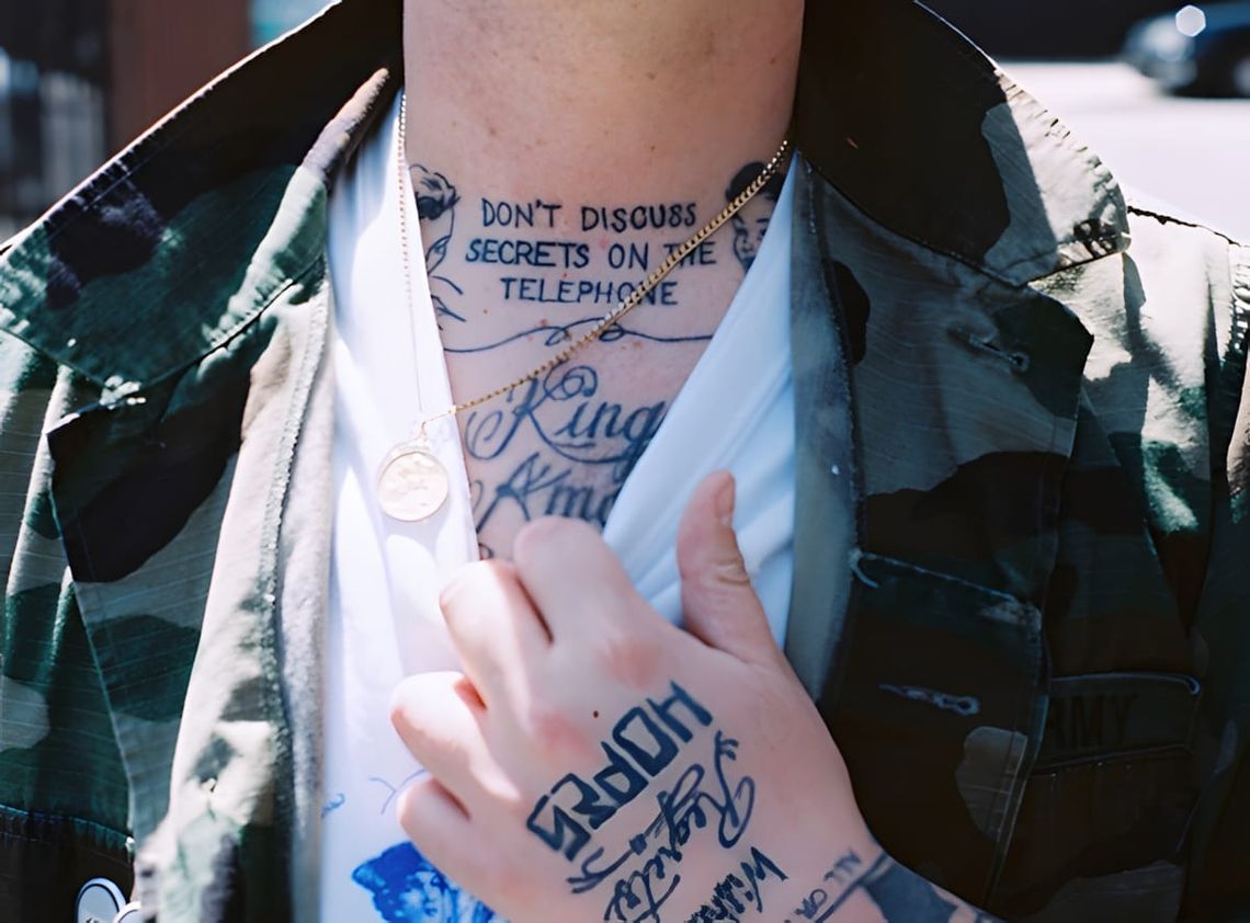 Close-up street photo of a tattooed person's chest and neck; neck reads "Don't Discuss Secrets on the Telephone" in block-let