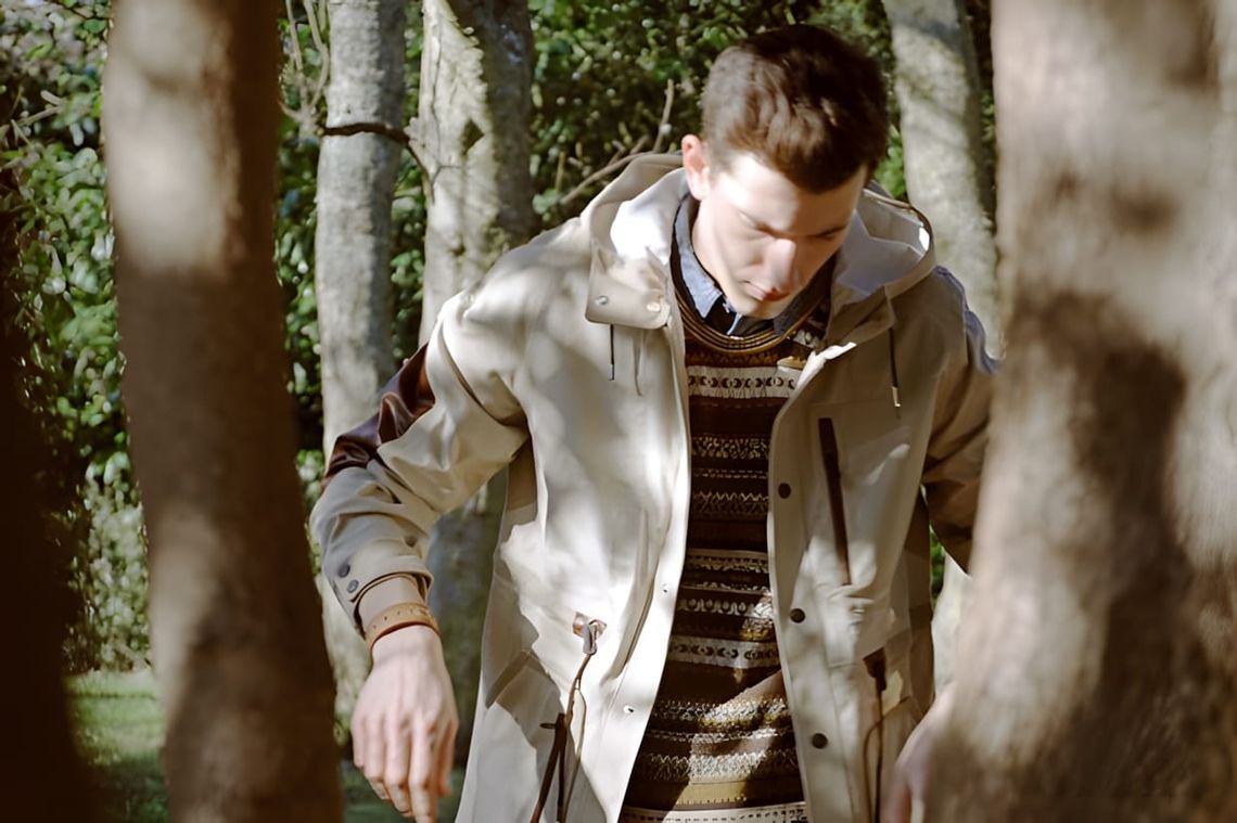 Male model in a forest, wearing a sand-coloured hooded drawstring shell jacket over a brown and olive geometric fair-isle kni