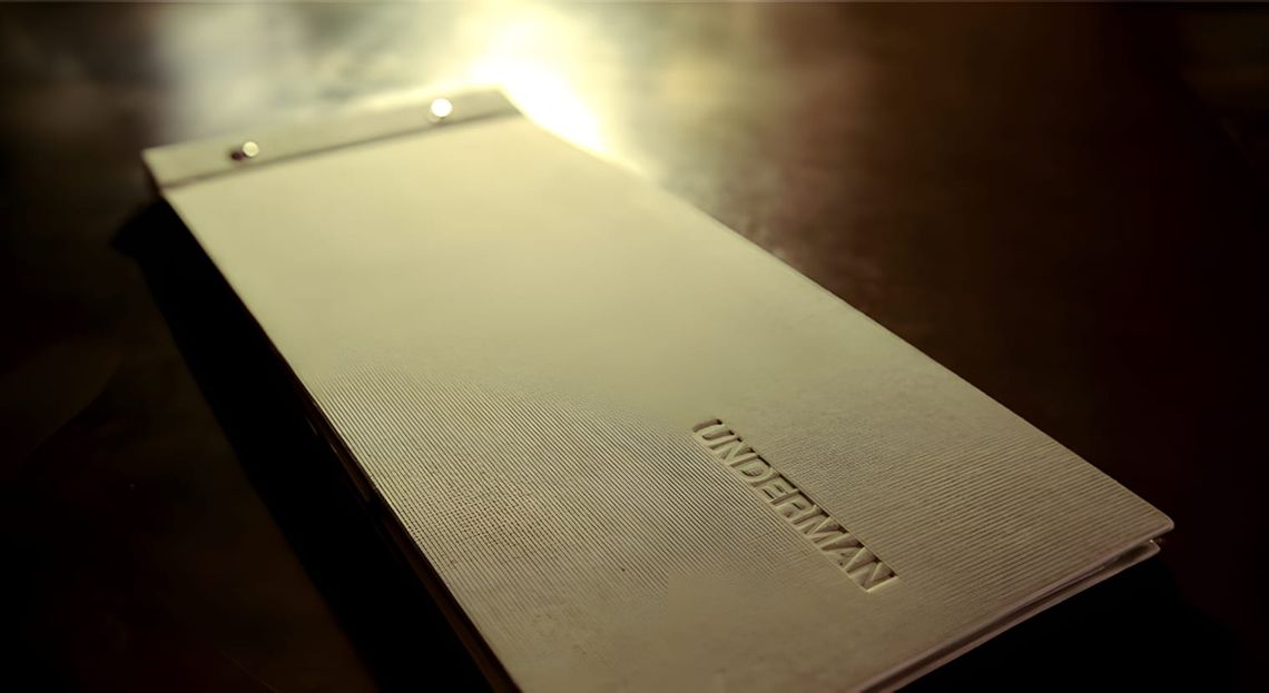 Landscape-format hardcover book lying flat on a dark surface; off-white linen-textured cover is debossed with the word "UNDER
