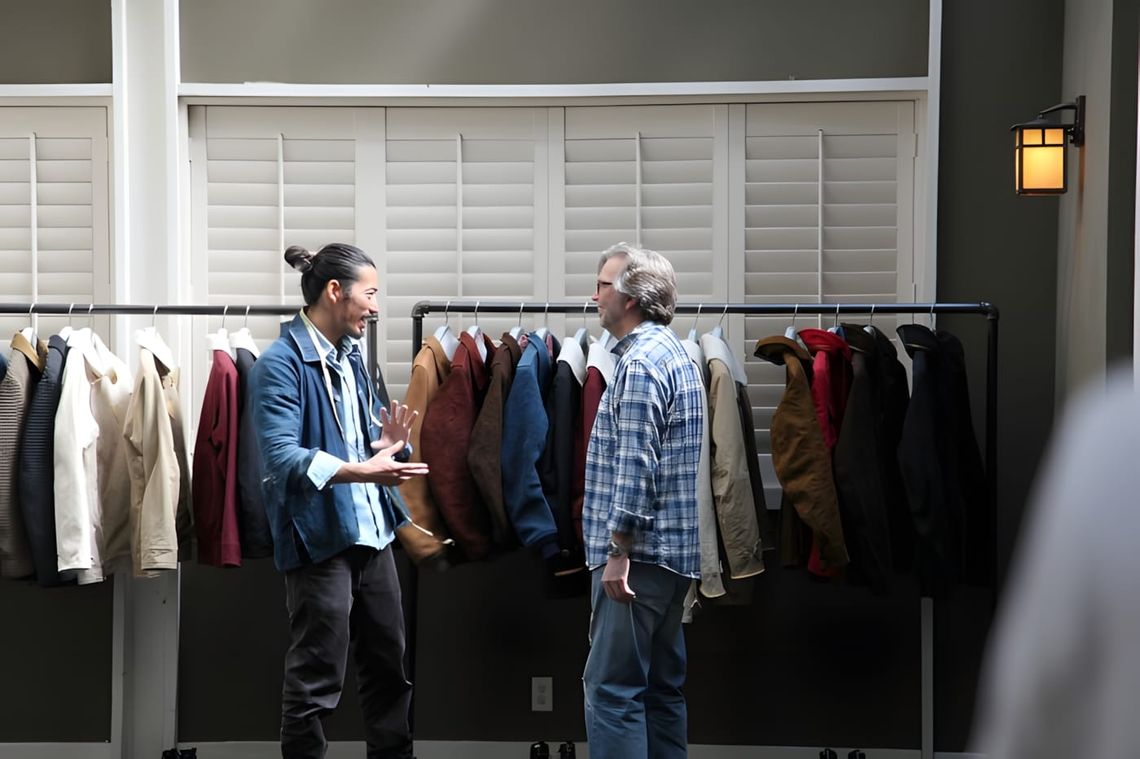 Two attendees talking in front of a clothing rack holding Visvim jackets and coats in navy, burgundy, tan, and brown at the V