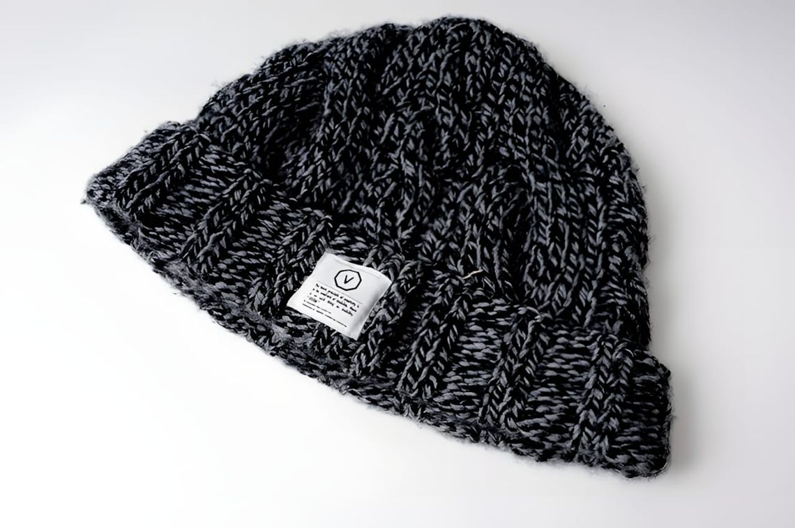 Visvim chunky cable/braid-knit cuffed beanie in marled grey and black wool, laid flat on a white surface, with a small white 