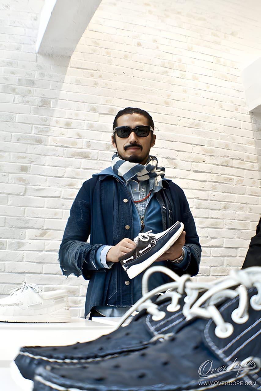 Hiroki Nakamura — top-knot, beard, sunglasses, denim jacket and plaid scarf — holds a pair of indigo navy visvim boat shoes w