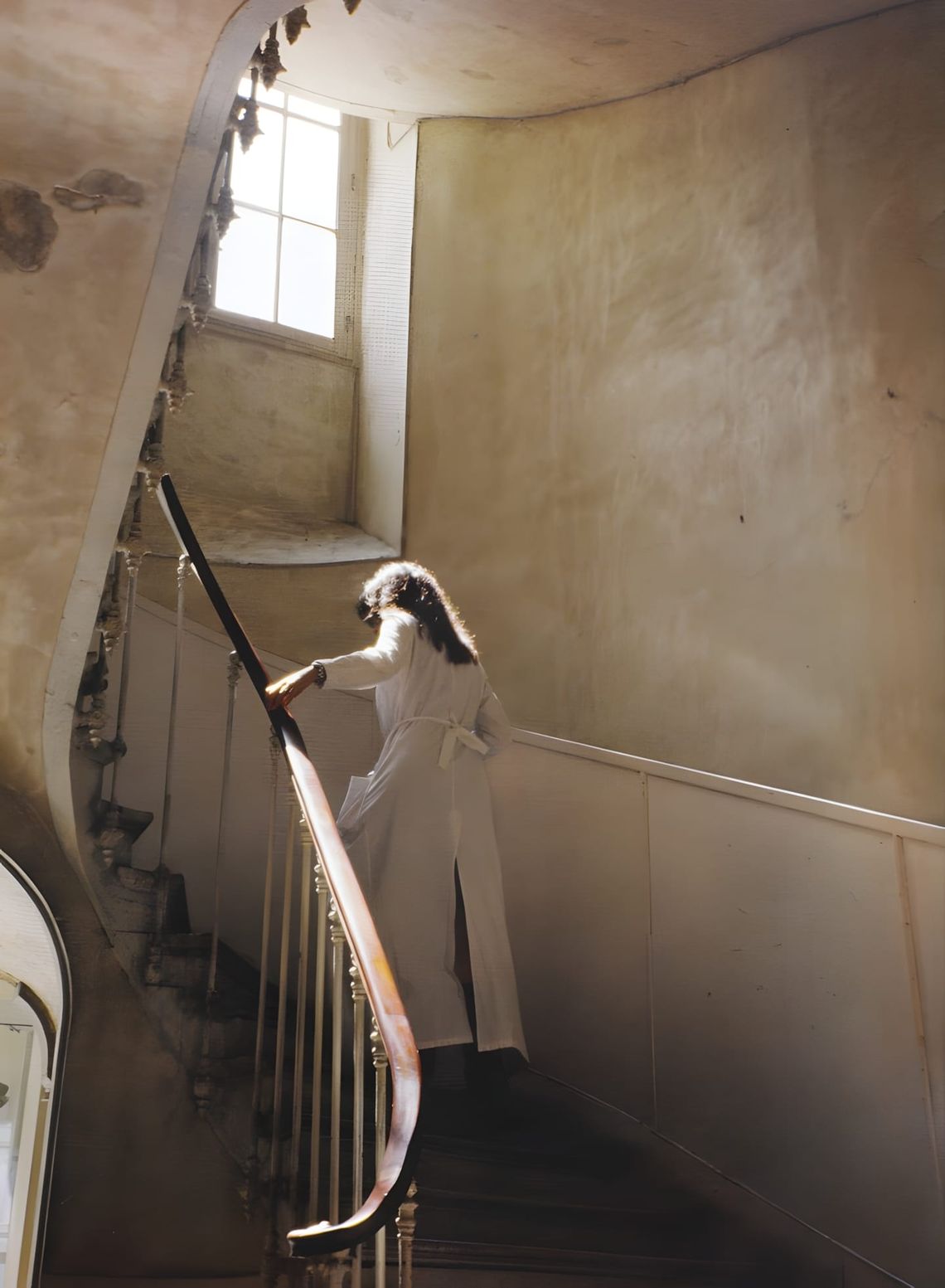 A figure in a white belted lab coat descends a curved wooden staircase in the dilapidated plaster interior of Maison Martin M