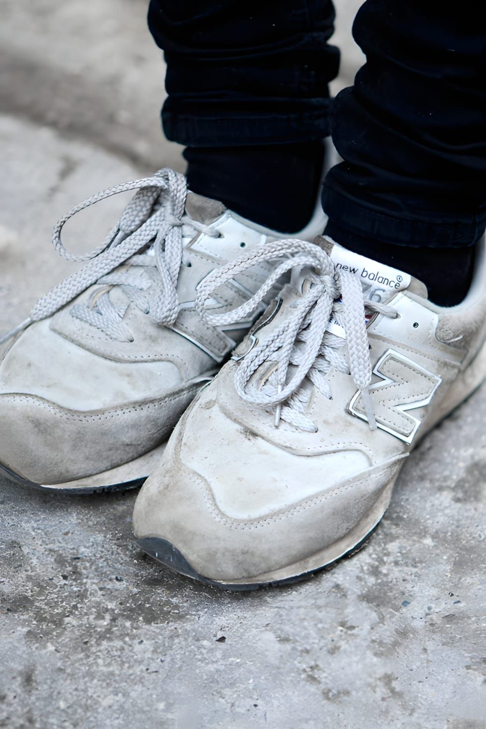 Worn pair of New Balance 574 grey suede sneakers on concrete, shot from above at foot level, showing heavily used light grey/