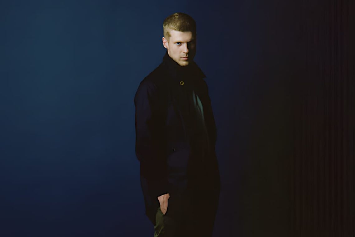 Male model in a long navy nanamica GORE-TEX Balmacaan coat, studio shot against a dark blue gradient background