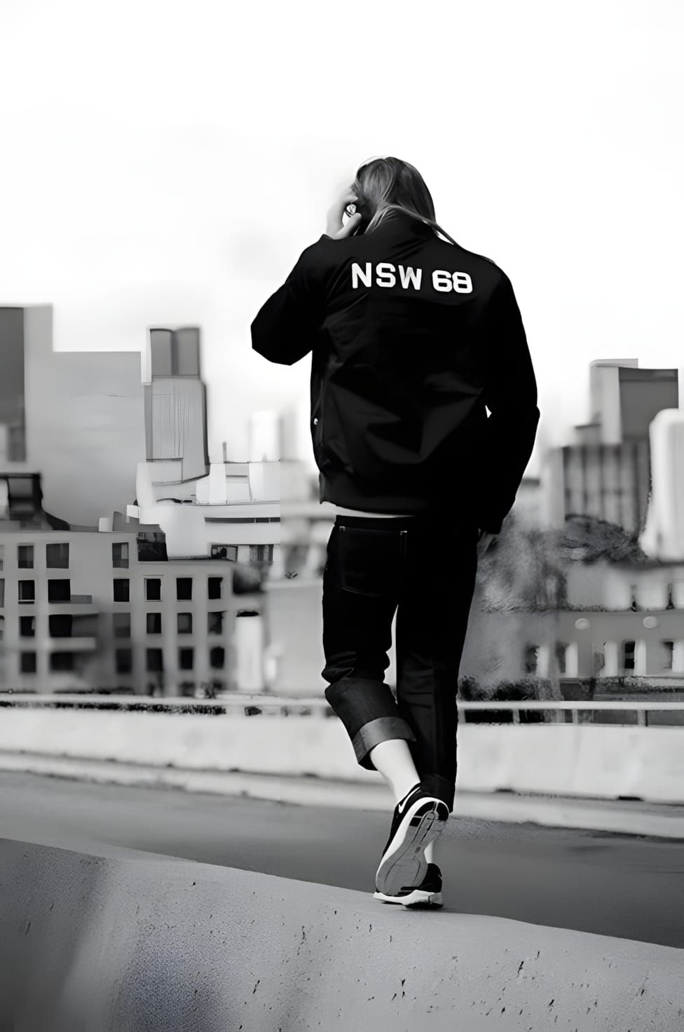 Figure on a rooftop from behind wearing a black Nike NSW 68 bomber jacket and dark jeans, city skyline behind.