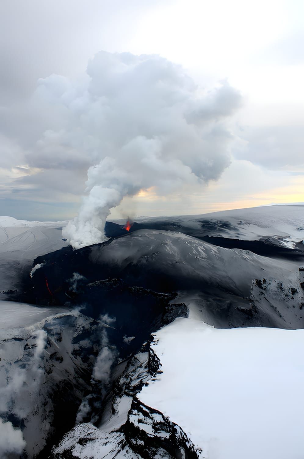 Aerial view of Plosky Tolbachik eruption, Kamchatka — dark lava field with glowing vent and billowing ash plume.
