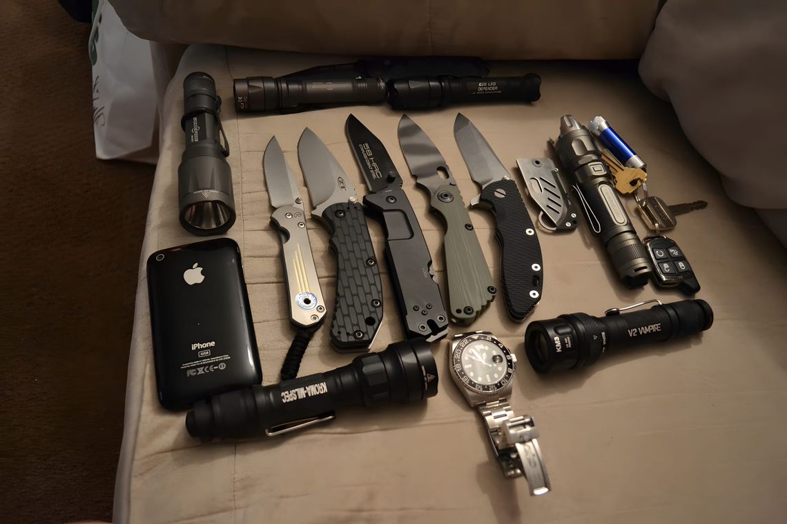 EDC flat-lay on beige fabric: five open folding knives, SureFire E2D Defender, V2 Vampire, KM3, iPhone 3G, and GMT watch.