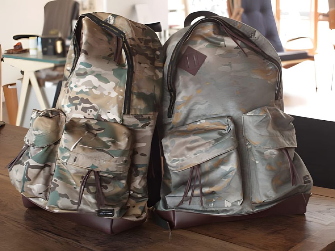 Two master-piece MSPC camo backpacks on a wooden table — MultiCam left, grey-green camo right, both with burgundy leather bas