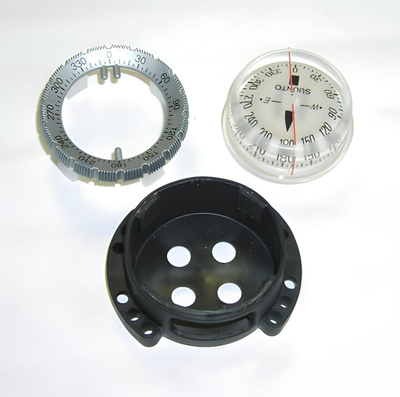 Suunto SK-8 compass disassembled: grey bezel ring, clear capsule with red needle, and black bungee mount bracket.