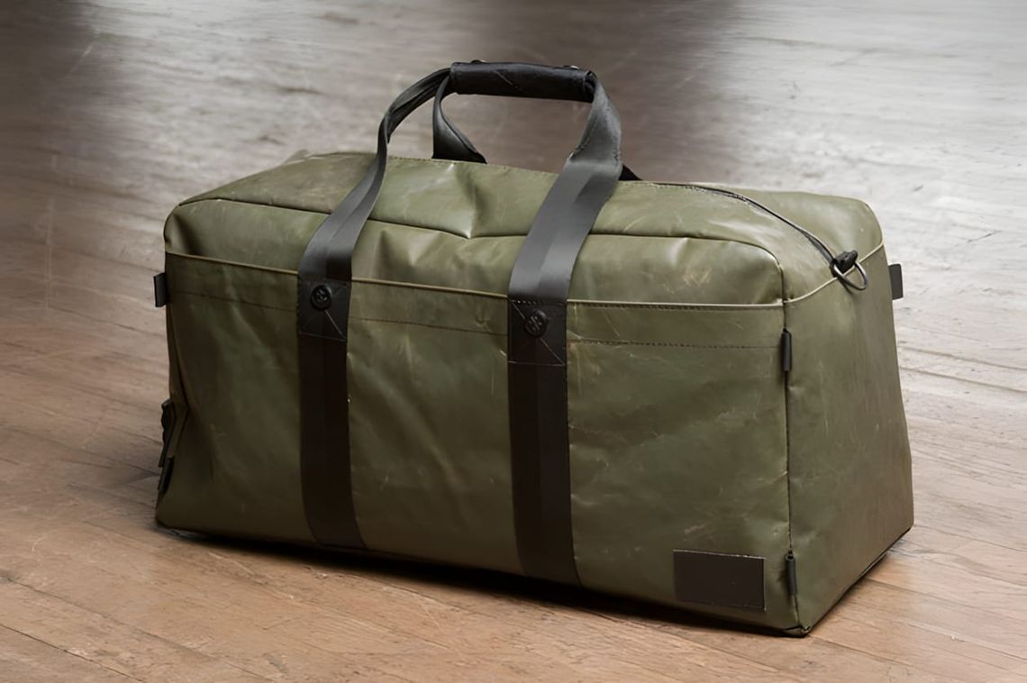 Killspencer duffle in olive drab duck canvas with dark leather handles and square leather patch, on wood floor.