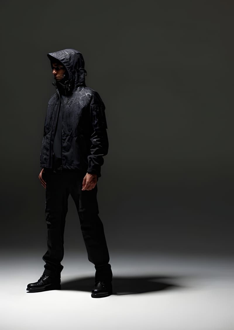 Model wearing Vollebak Full Metal Jacket copper edition — hooded dark copper-fibre shell, bellows pockets, spotlight on grey 