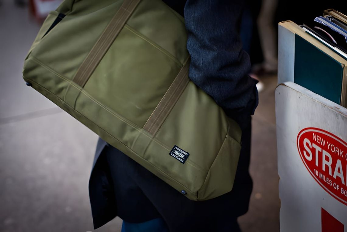 Olive Cordura Monocle x Porter Boston Bag with brown webbing straps carried under the arm on a New York street, Strand Bookst