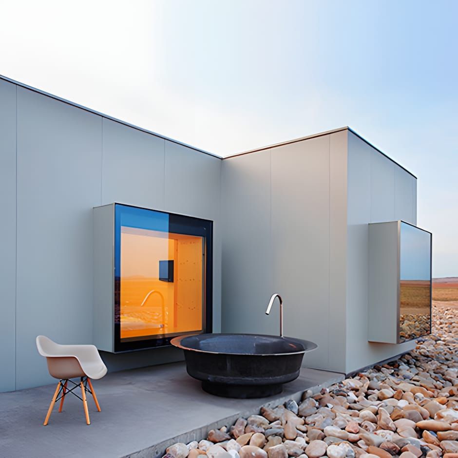 Grey metal cube hotel room with amber-lit picture window, freestanding black stone bath, and Eames DAW chair on desert terrac