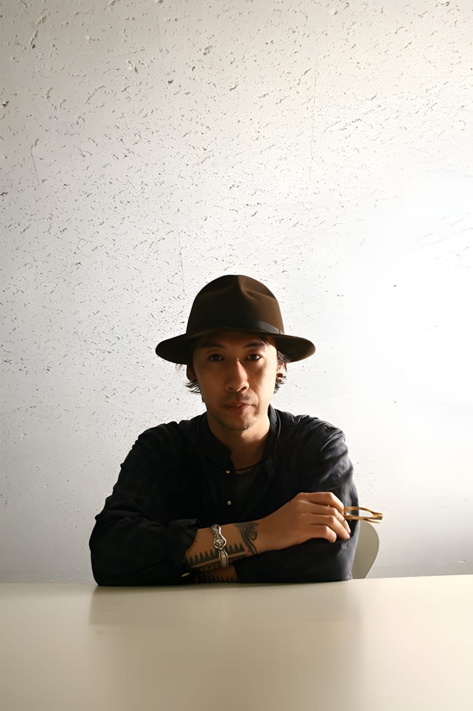 Jun Takahashi seated at a white table, wearing all black and a dark wide-brim felt hat, forearm tattoos visible, holding glas