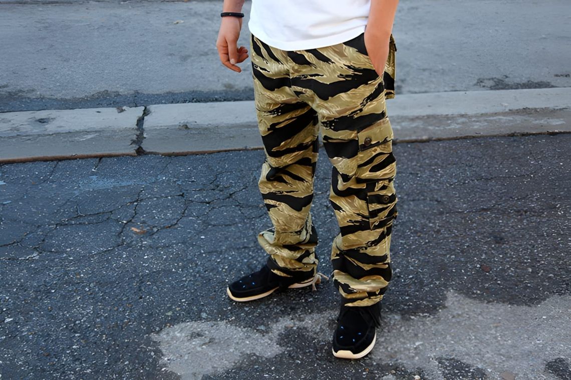 Man in gold and black tiger stripe camo cargo pants with black Nike sneakers on a city sidewalk.