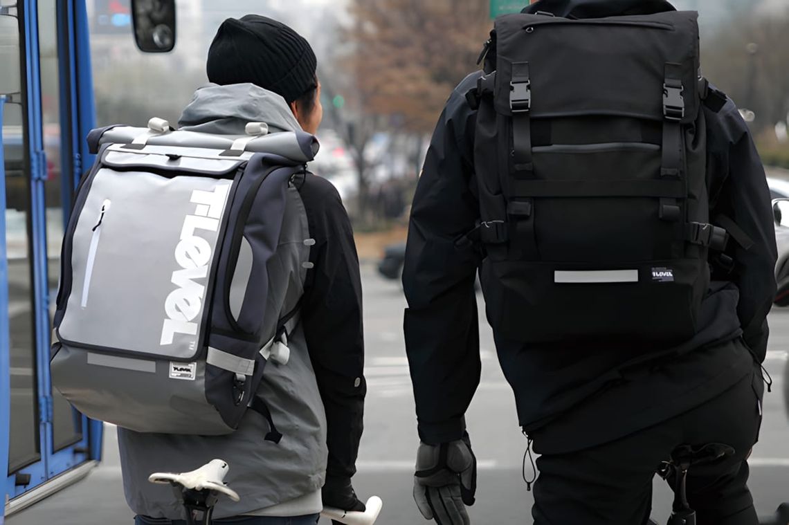 Two cyclists from behind wearing T-Level Infinity Rolltop backpacks in grey and black on an urban street.