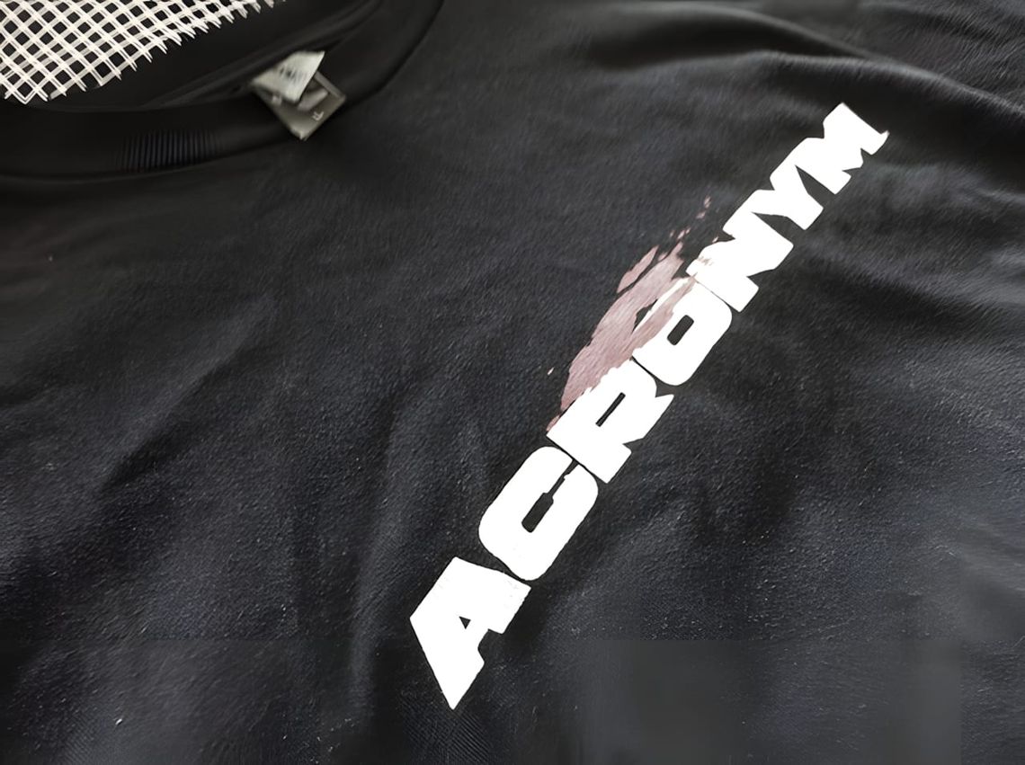 Black ACRONYM T-shirt with vertical white logo print and distressed ink-splash graphic on the chest.