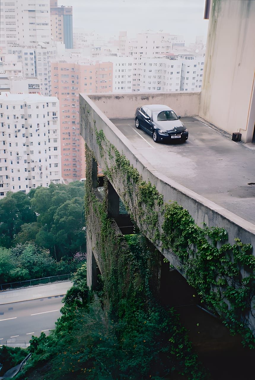 Overhead view of a dark navy BMW 3 Series E90 parked on the top level of a vine-covered brutalist concrete car park, with Hon