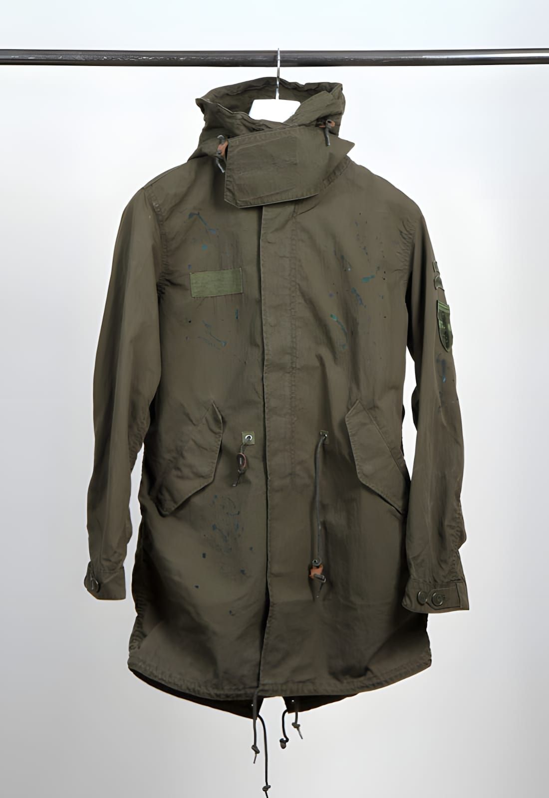 uniform experiment Damaged Mods Coat, an olive fishtail parka with badge embellishments and paint splatter detailing