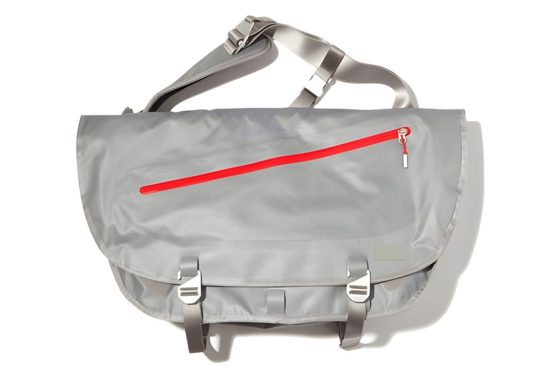 Nike Cascade Messenger bag in silver-grey nylon with diagonal red zipper and silver buckle hardware, product shot on white.