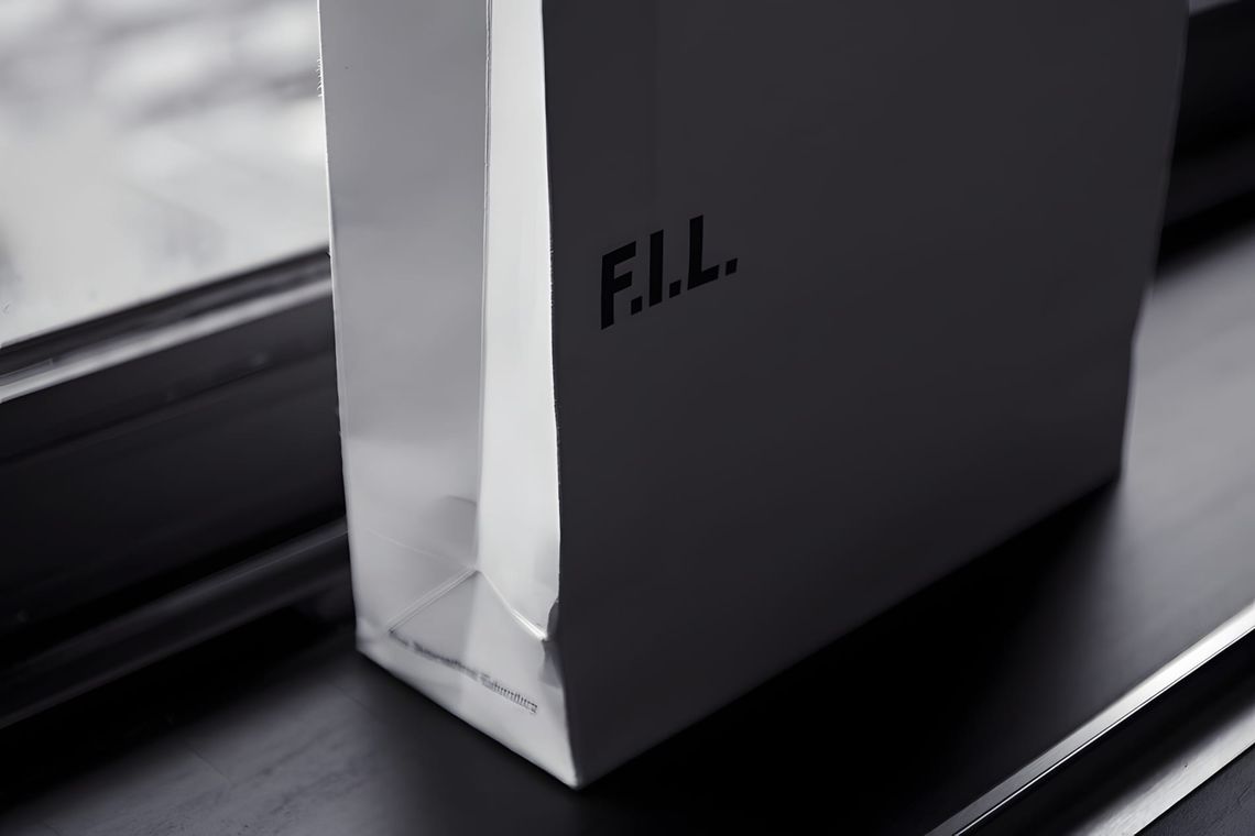 Matte black debossed F.I.L. gift box beside a white paper shopping bag on a dark surface.