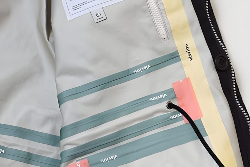 Interior lining of a visvim jacket showing teal stripe seam tape printed with 'visvim' text repeatedly, interior zip and snap