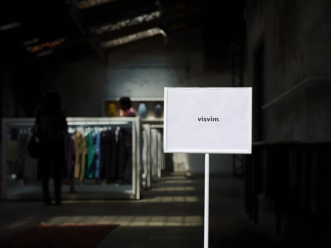 Interior of a Visvim showroom: a white placard bearing the 'visvim' logo stands on a thin metal pole in the foreground; in th