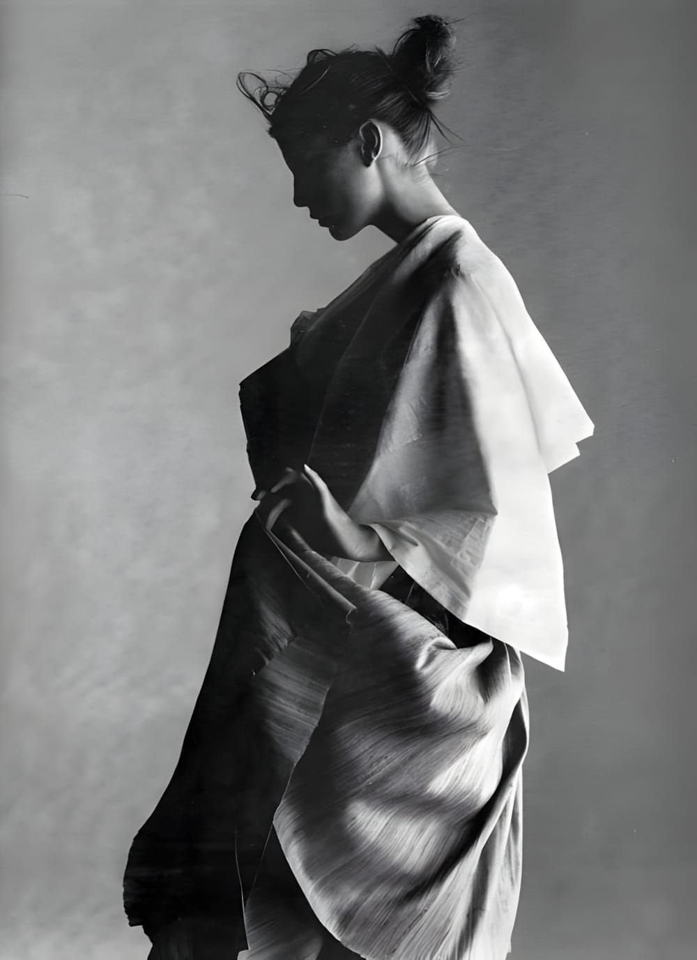 Black-and-white studio photograph of a female model in side profile, wearing a voluminous pale wide-sleeved wrap top and a de