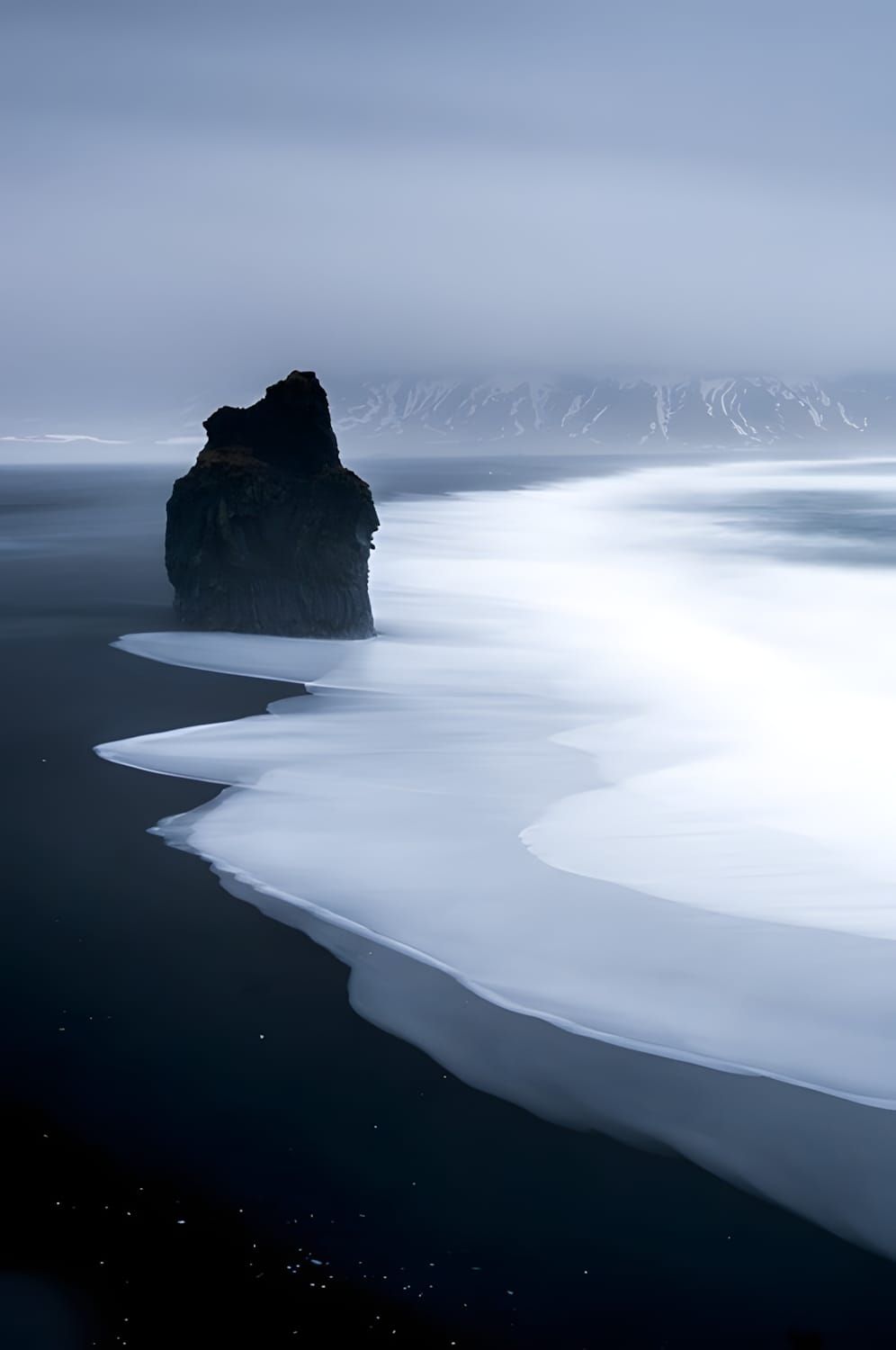 Lone basalt sea stack at Reynisfjara black-sand beach, Iceland; long-exposure surf curves white around it, misty mountains be