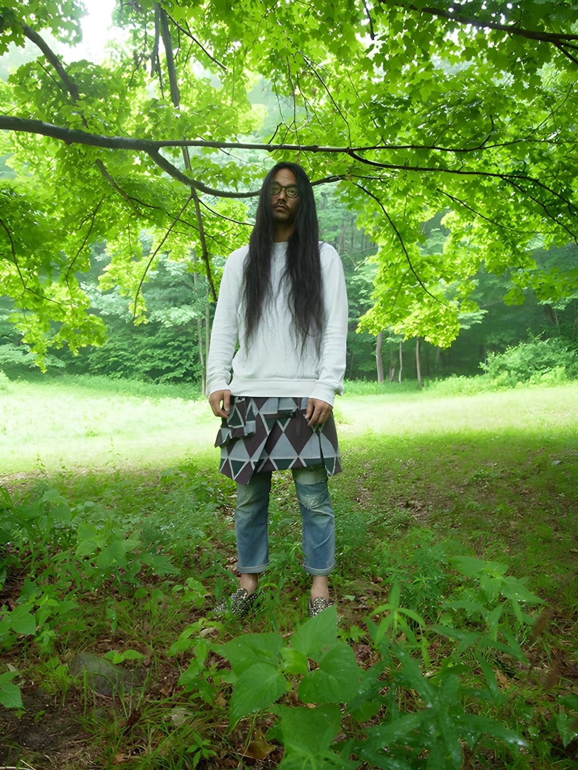 Long-haired model standing in New Zealand forest wearing white sweatshirt and Kapital diamond-pattern wool wrap skirt over je