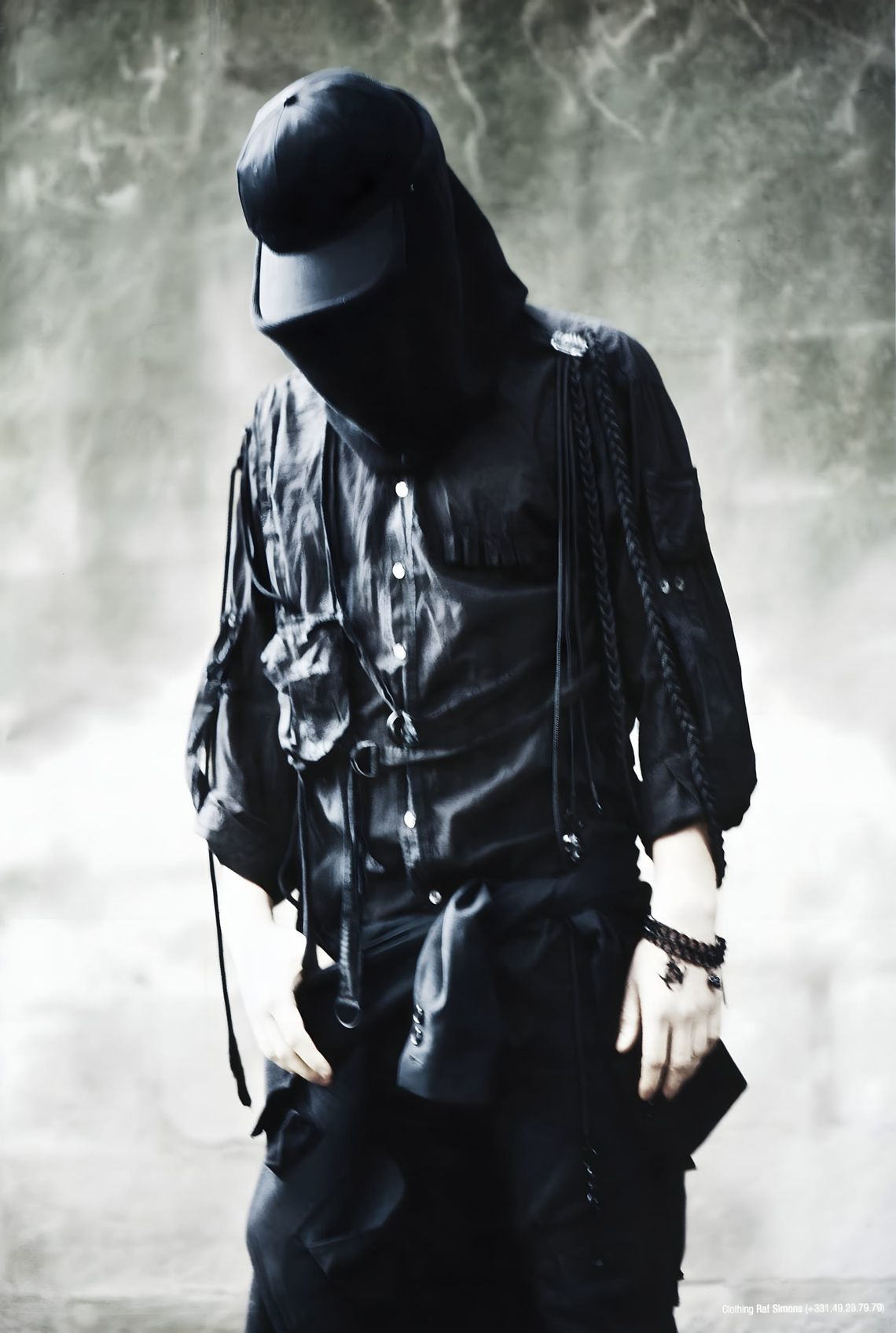 Model in all-black Raf Simons look: cap over hood, sheer utility shirt with D-rings and hanging straps, braided leather wrist