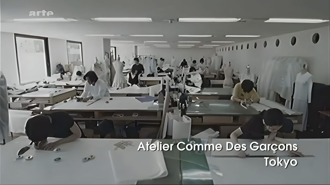 Black-and-white video still showing the Comme des Garcons Tokyo atelier; workers cut fabric at long tables beside dress forms
