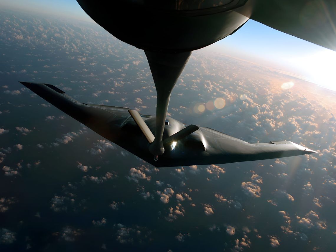 Northrop B-2 Spirit flying-wing stealth bomber receiving in-flight refueling above clouds, viewed from below the tanker.