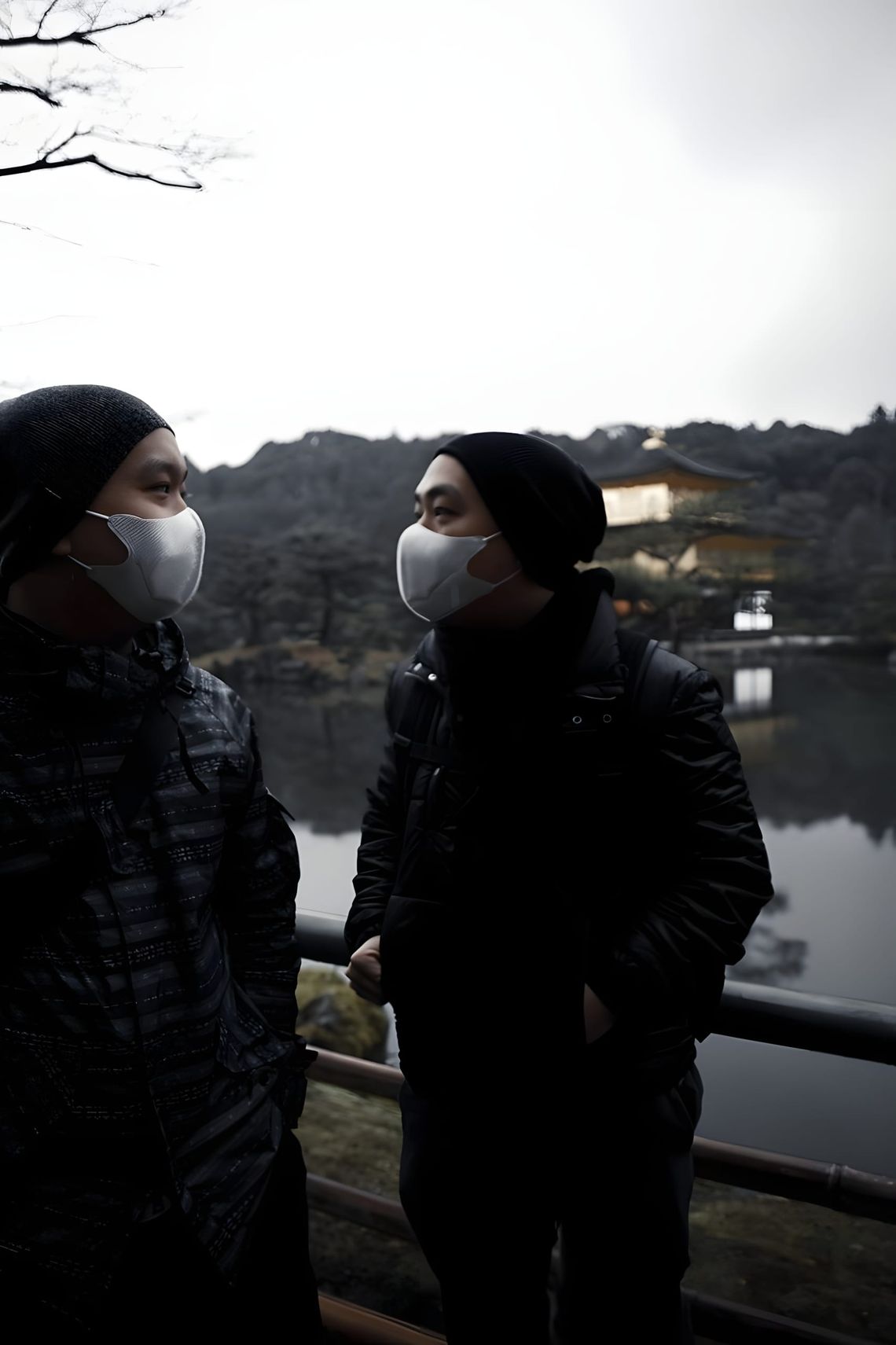 Two men wearing surgical face masks and beanies stand at Kinkaku-ji (Golden Pavilion), Kyoto; the figure on the left wears th
