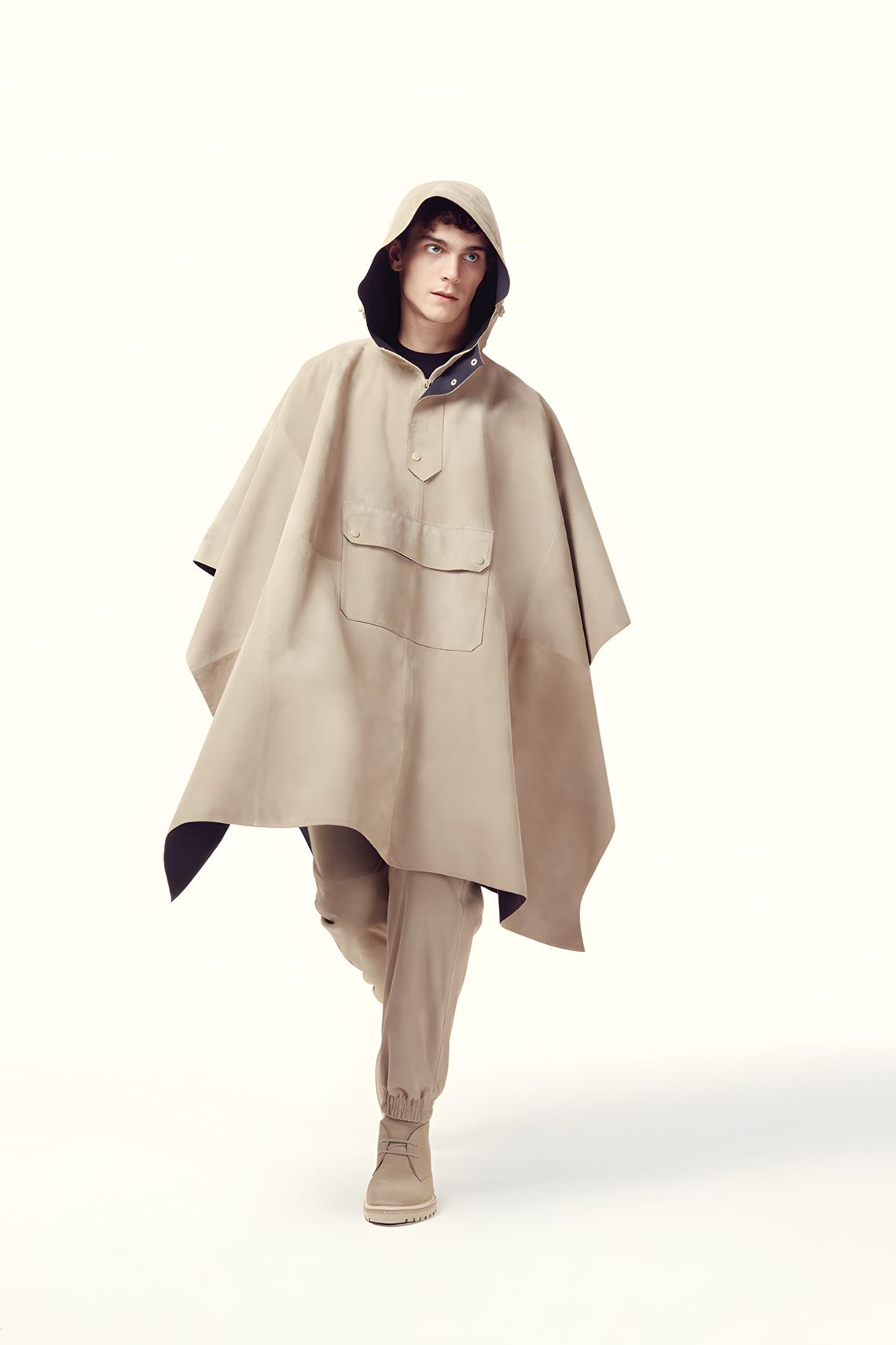 Male model on a clean white runway wearing a tan hooded cape-poncho with a utility chest pocket, matching tapered trousers, a