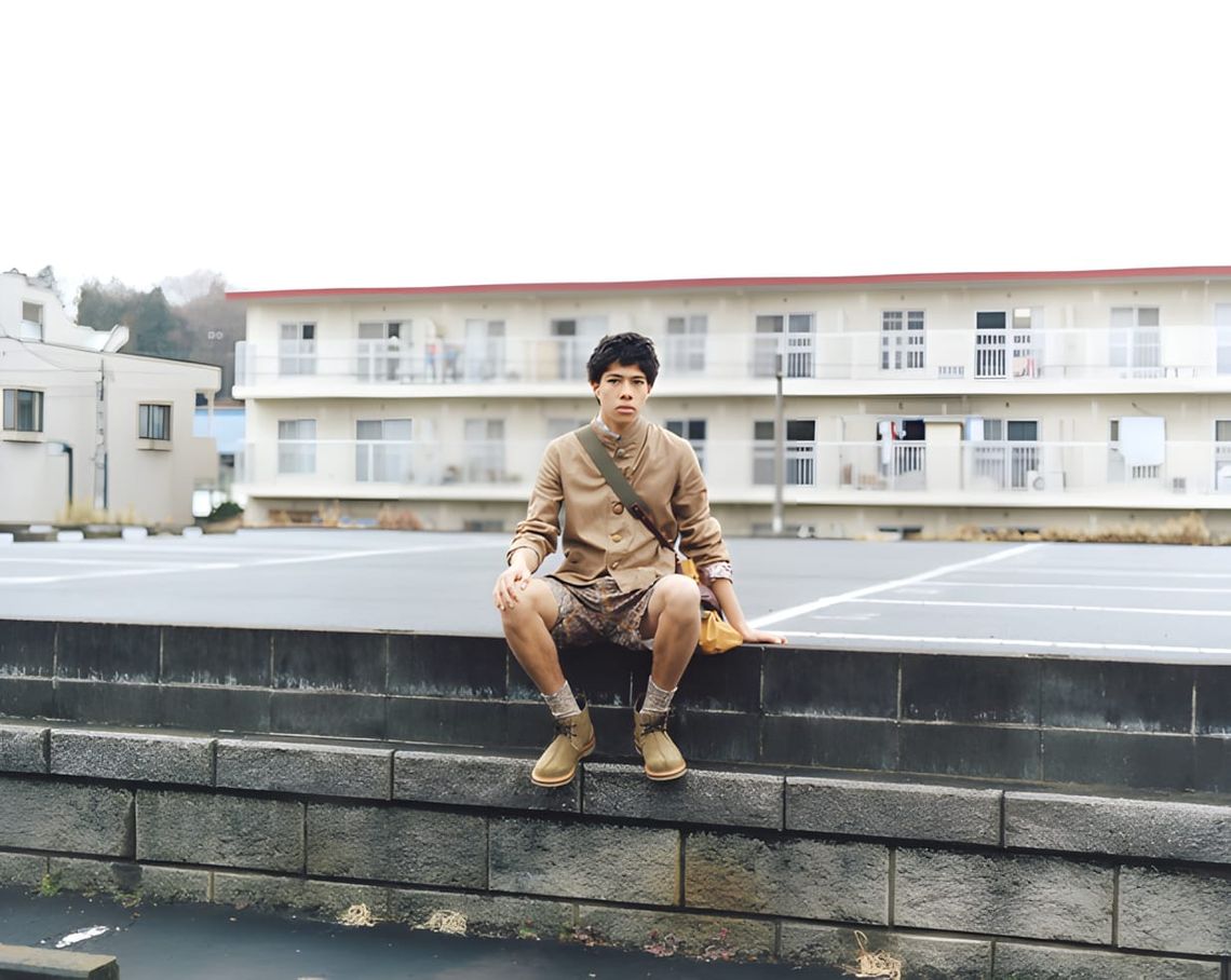 Young Japanese man in a beige button-up shirt, shorts, and desert boots sitting on a concrete garden wall in front of a low-r